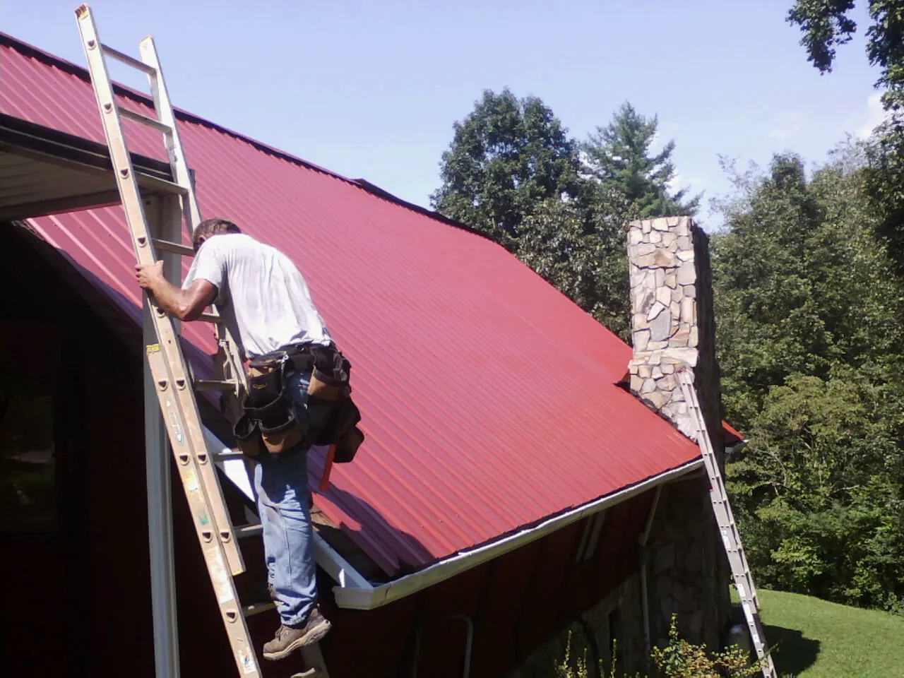 Roofer on a ladder beside a red metal roof and stone chimney — Soffit & Fascia Repair by Roofing Brothers in Cosmopolis